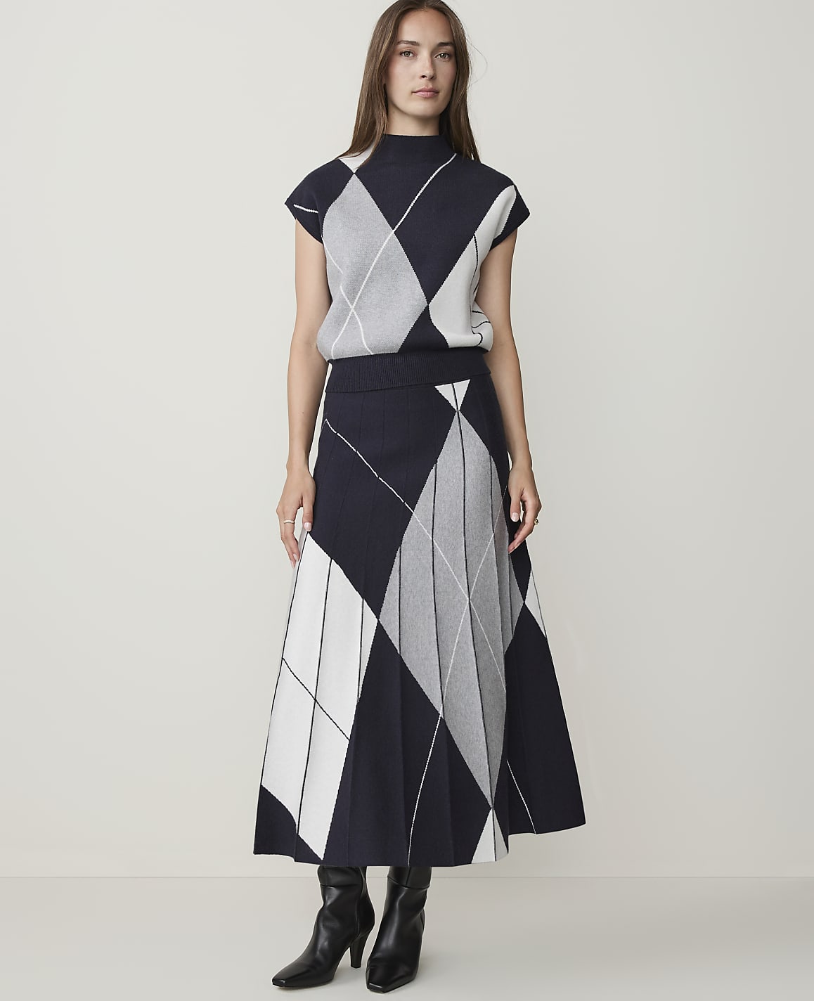A woman in a navy, white, and gray argyle knit top and pleated skirt stands against a plain, light gray background.