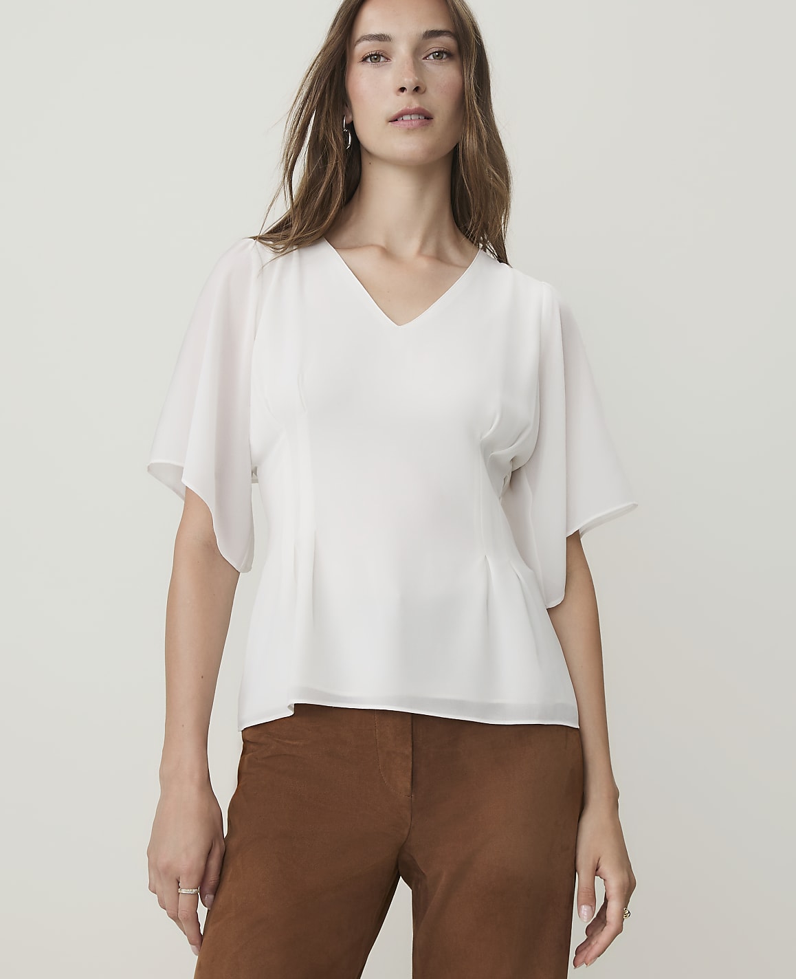 A woman in a white v-neck blouse and brown pants stands against a plain, light gray background.
