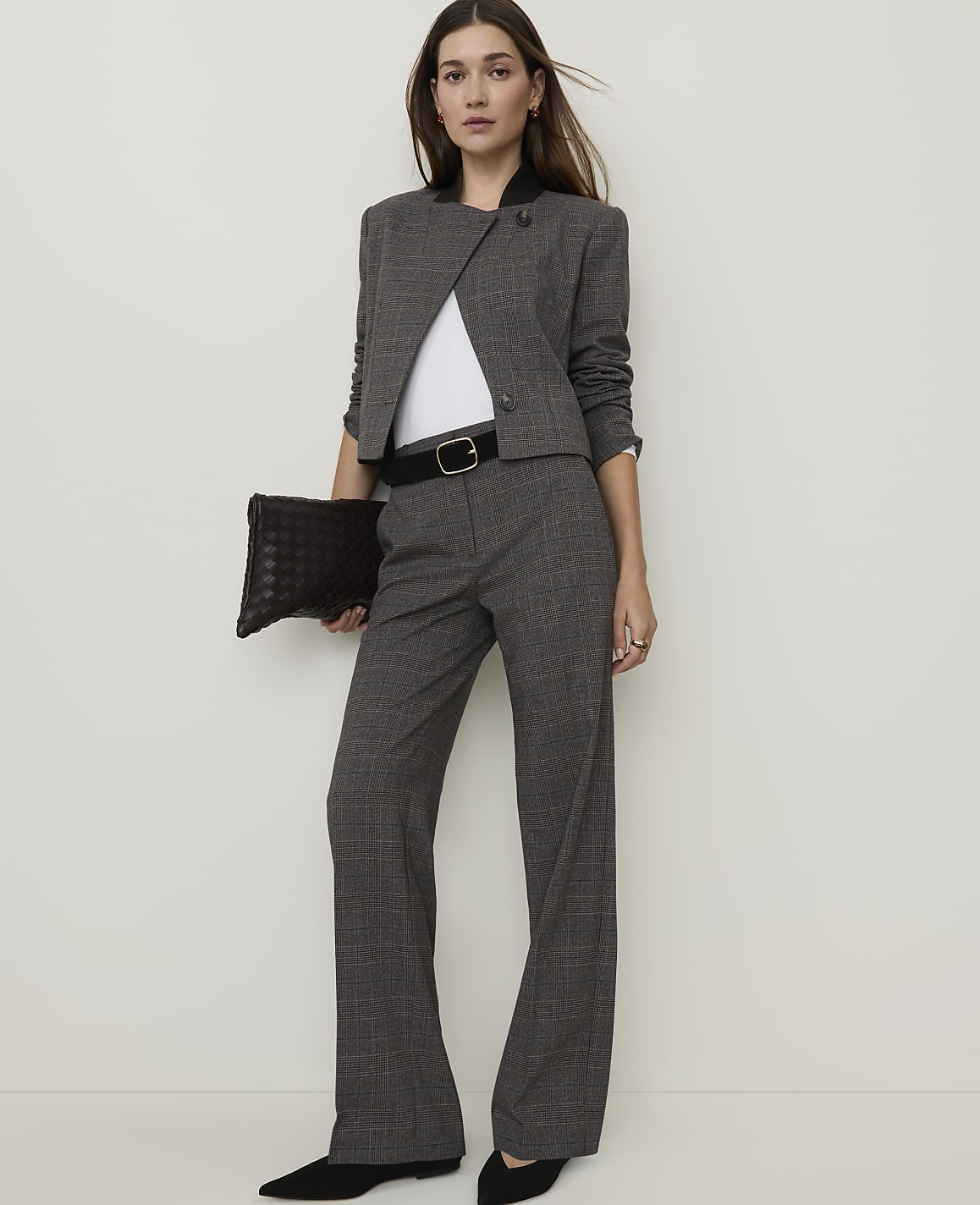 A woman in a grey plaid suit and black heels stands holding a dark woven clutch against a plain white background.