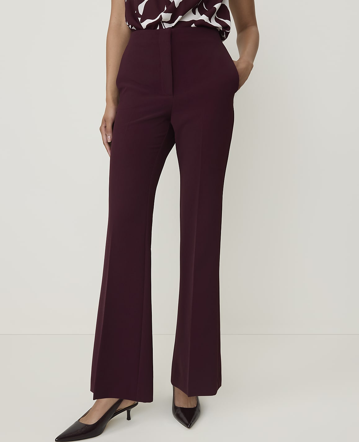 A person in maroon flared trousers and black pointed-toe heels stands against a plain white background.