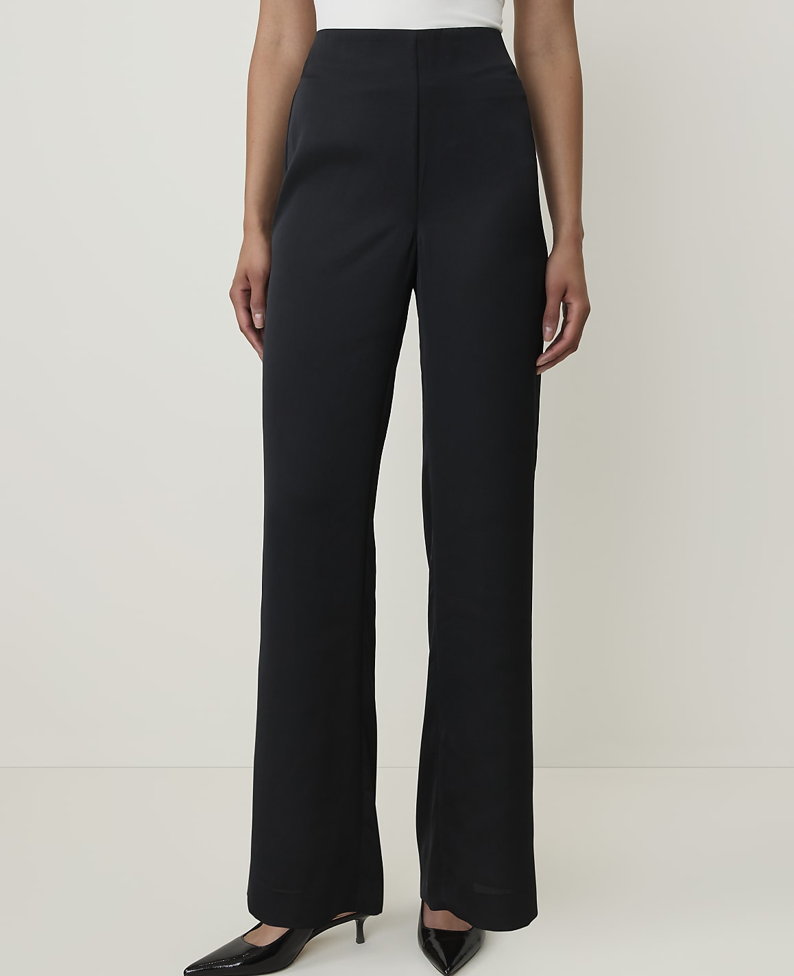 A person stands in black wide-leg pants and black pointed-toe heels against a plain white background.