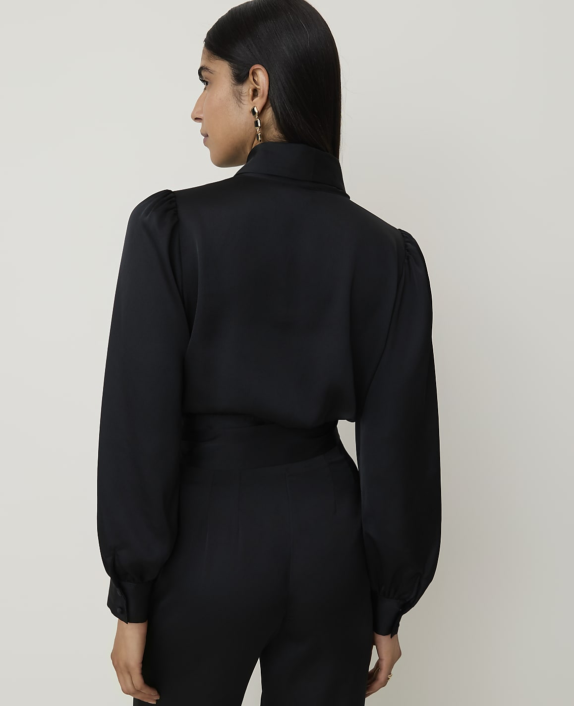 A woman in a black satin blouse with puffed sleeves and high-waisted pants stands against a plain white background.