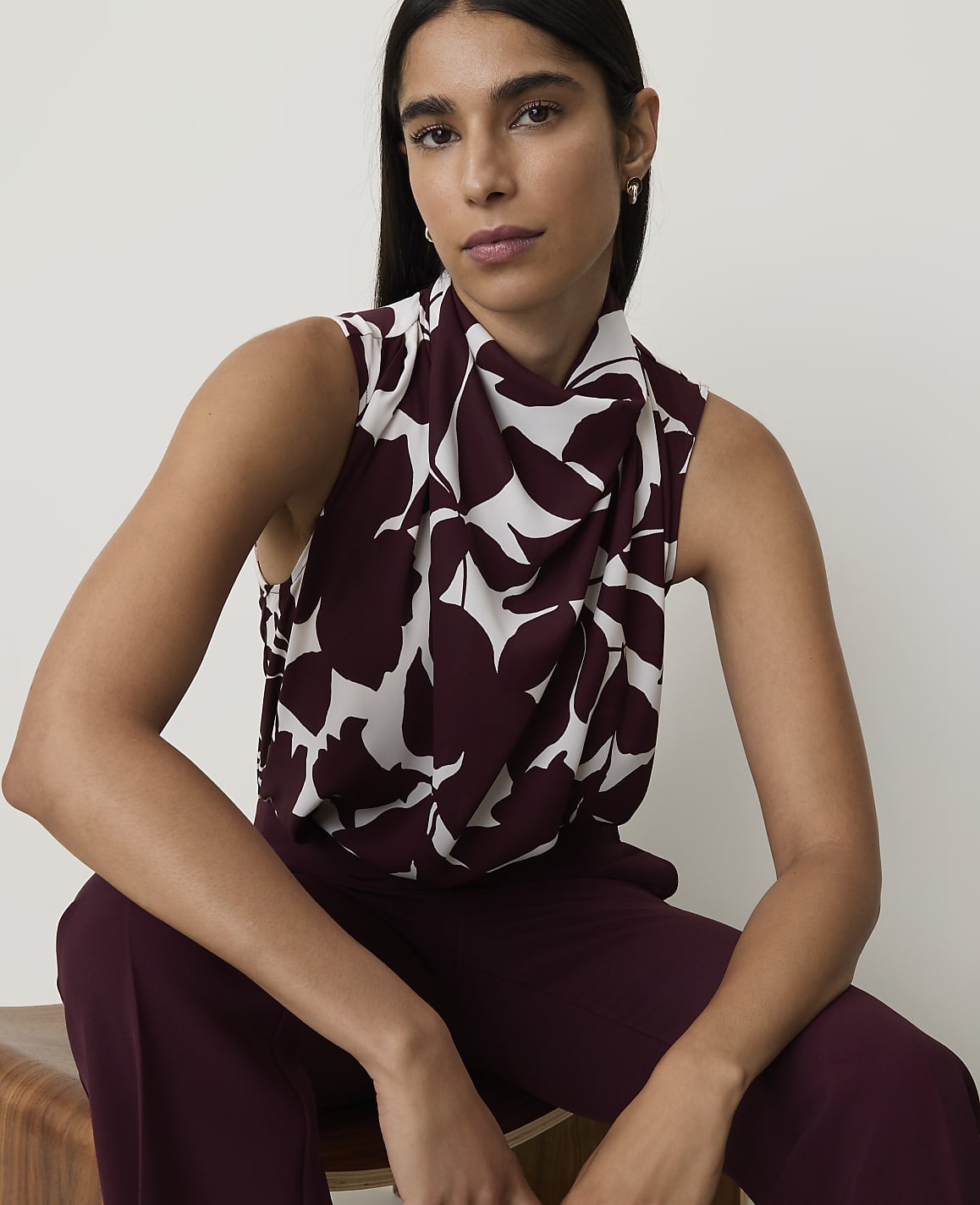 A woman in a patterned blouse and maroon pants sits in front of a plain white background.