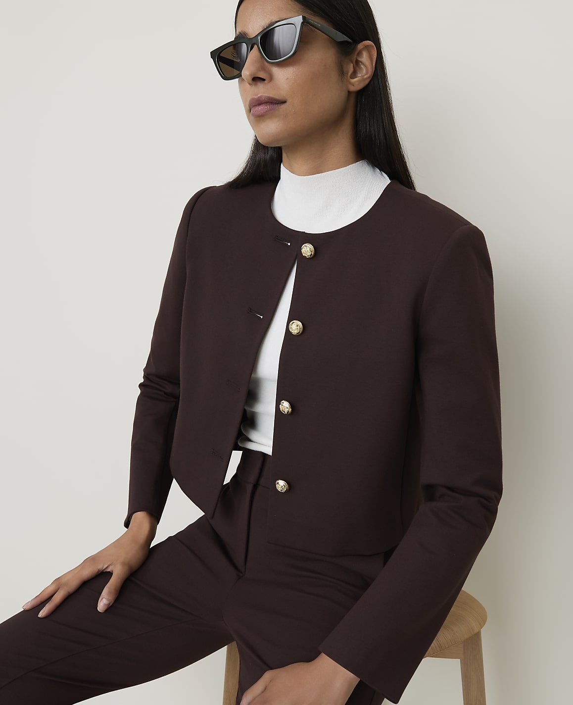 A woman in a brown jacket and white turtleneck poses against a plain background.