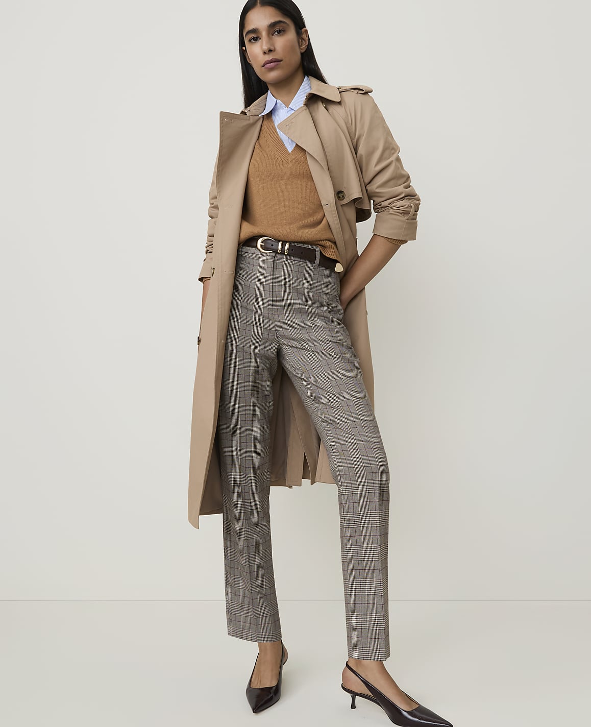 A woman in a trench coat, v-neck sweater, and plaid pants stands against a plain, light-colored background.