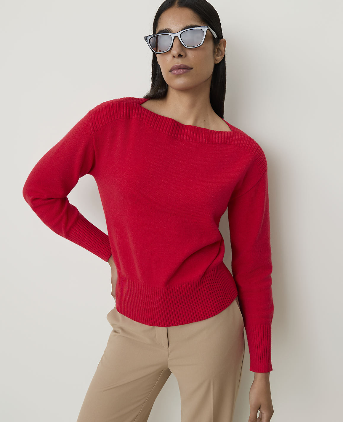 A woman in a red sweater and tan pants poses against a plain white background, wearing stylish sunglasses.