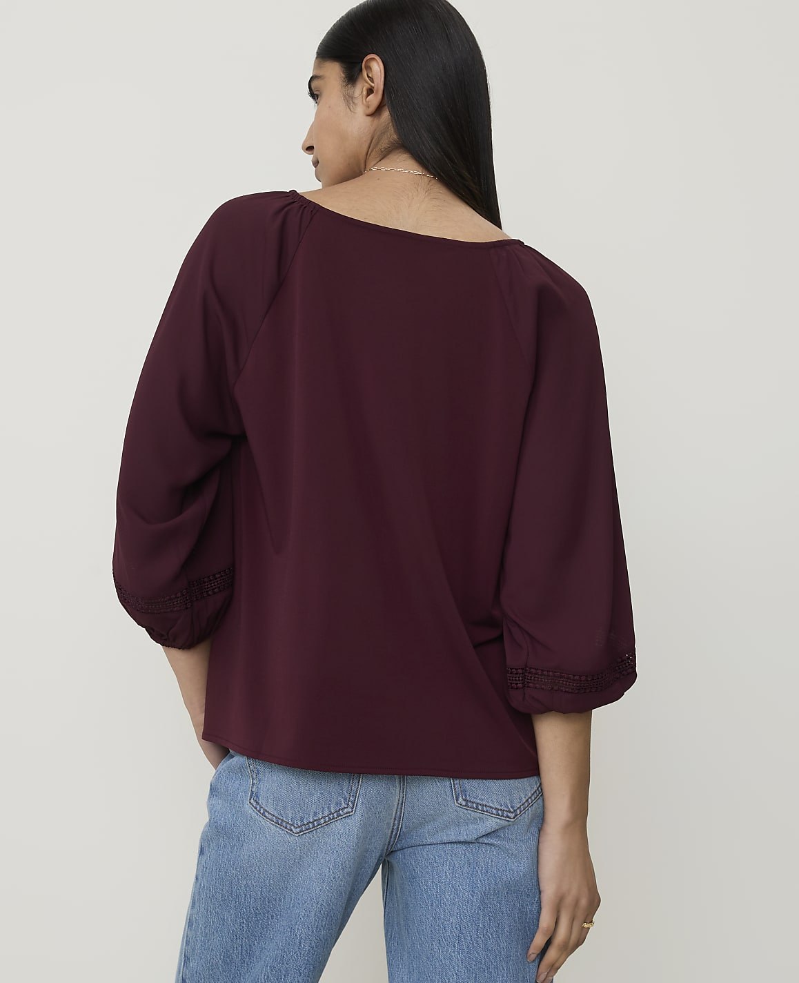 A woman in a maroon blouse and blue jeans stands against a plain white background.