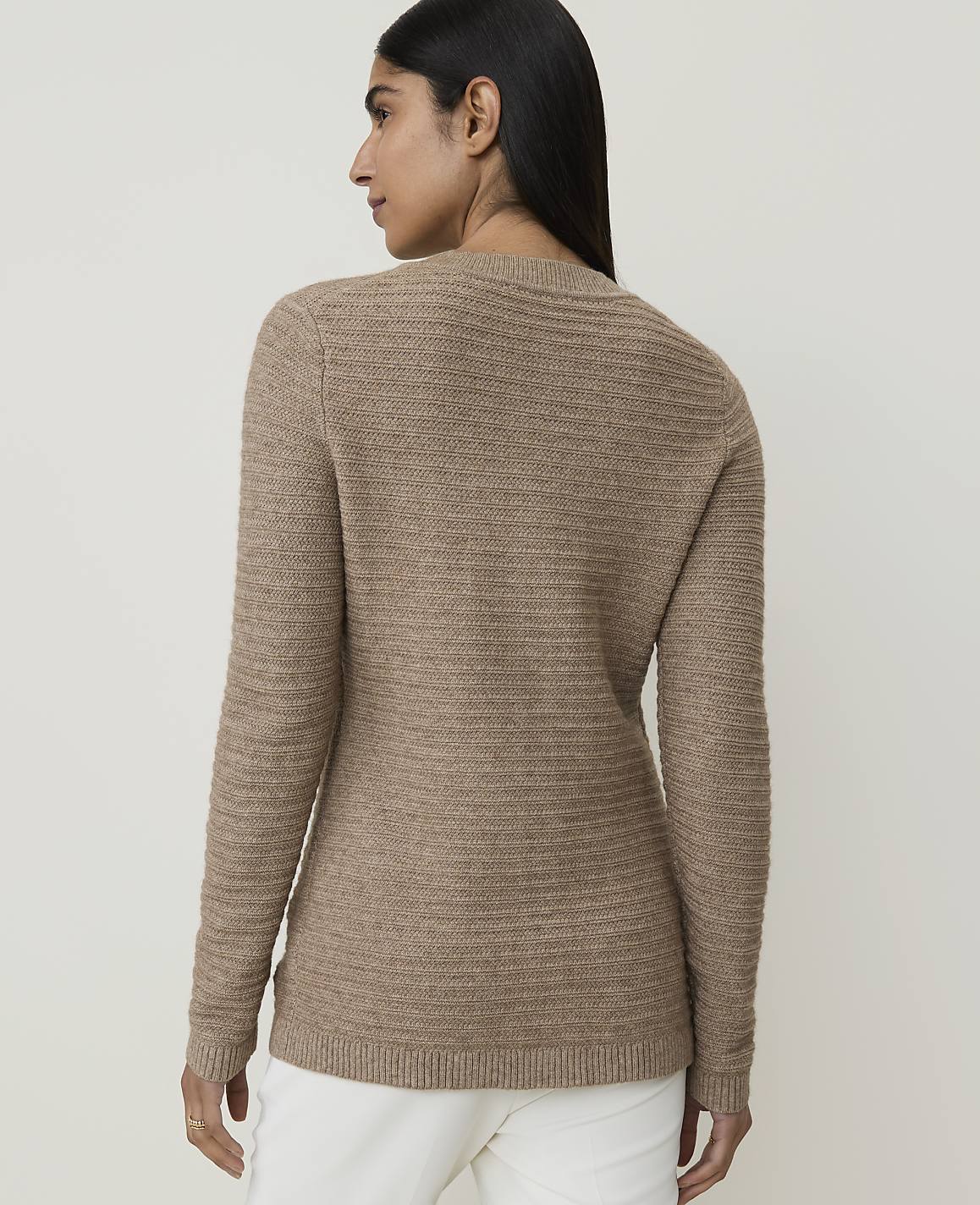 A woman in a brown textured sweater and white pants stands against a plain, light background.