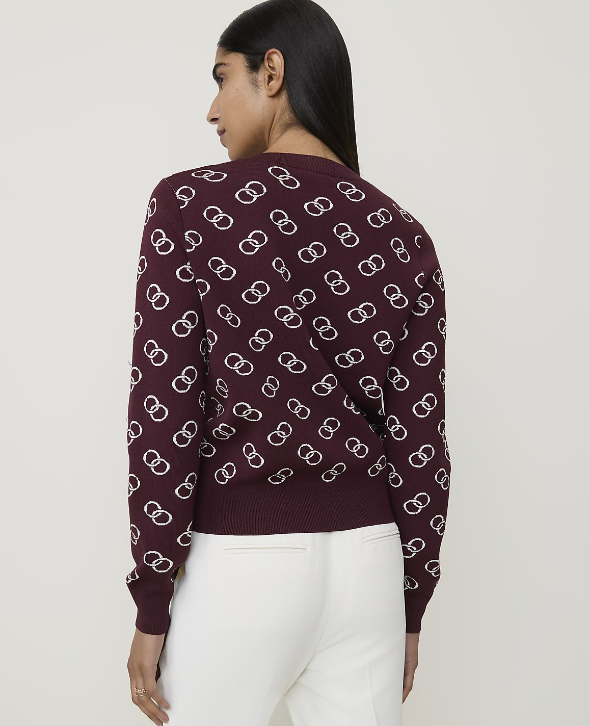 A woman in a maroon sweater with a white interlocking ring pattern stands against a plain background.