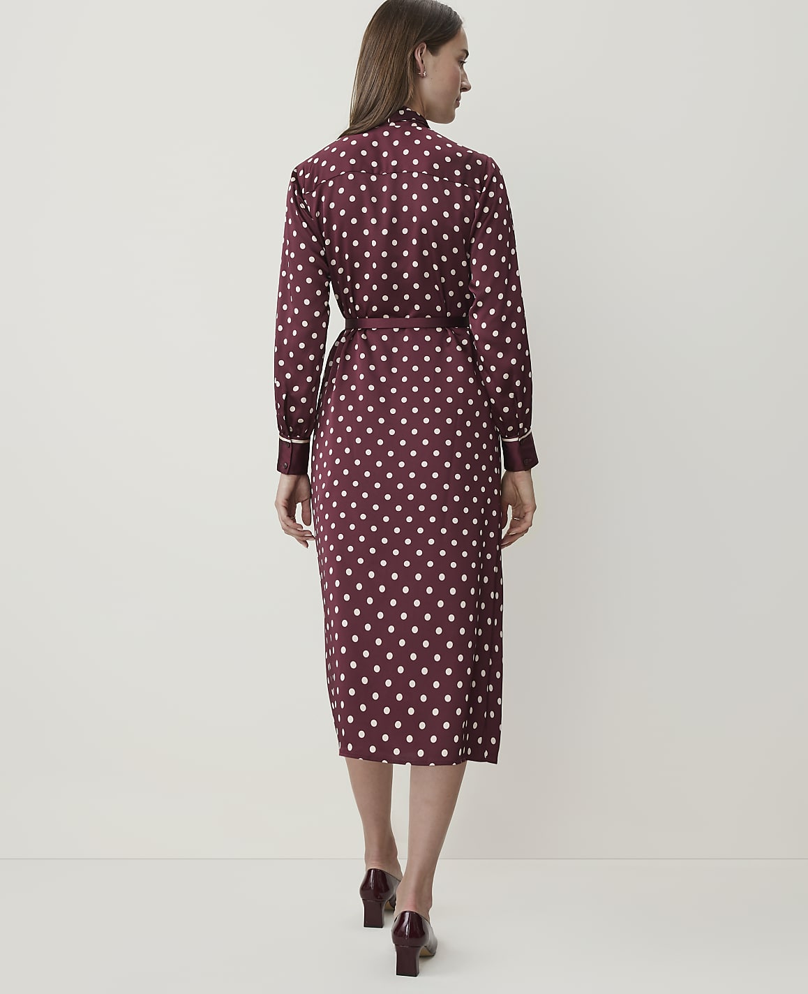 A woman in a burgundy polka dot dress stands against a plain white background.