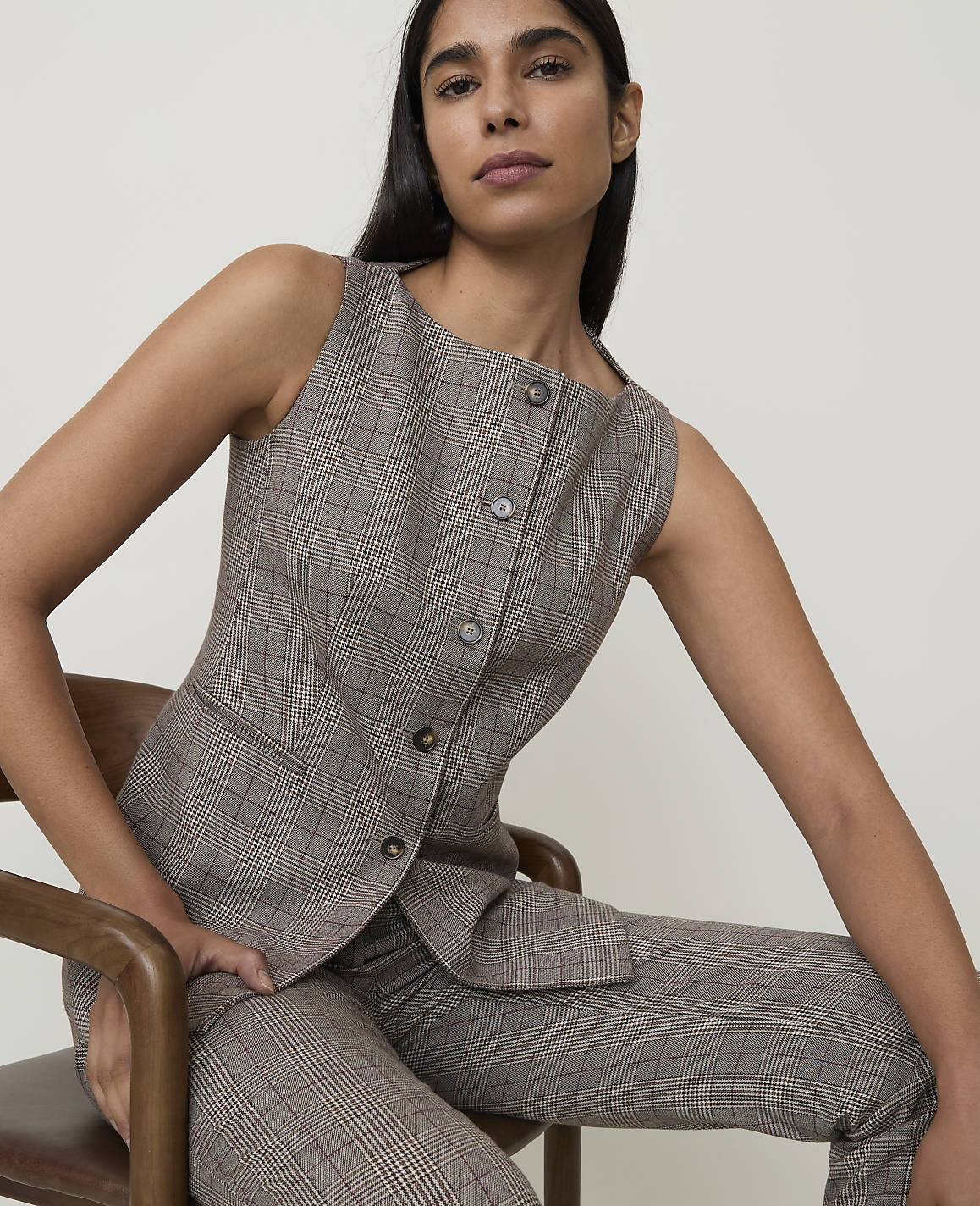 A woman in a plaid vest and pants sits on a wooden chair against a plain background.