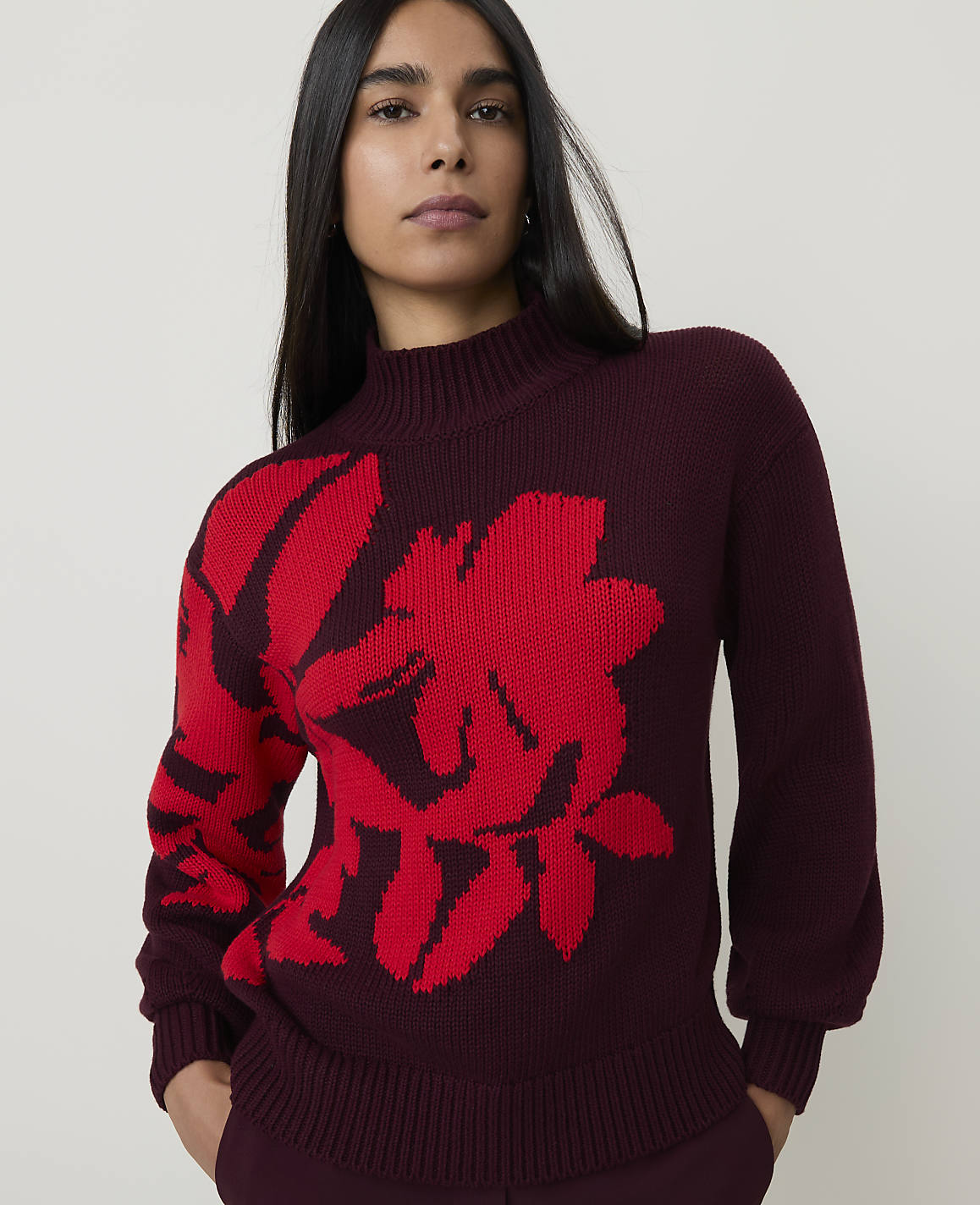 A woman in a maroon sweater with a large red floral design stands against a plain, light-colored background.