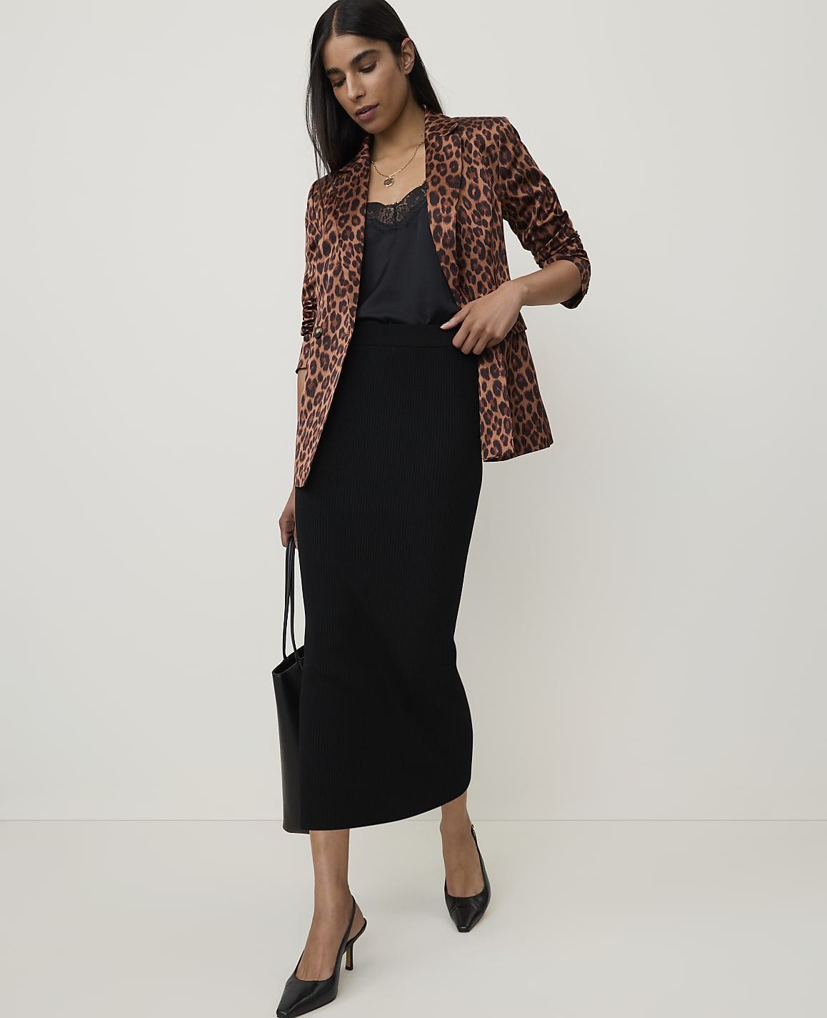 A woman in a leopard print blazer and black skirt stands against a plain, light background, holding a black tote bag.