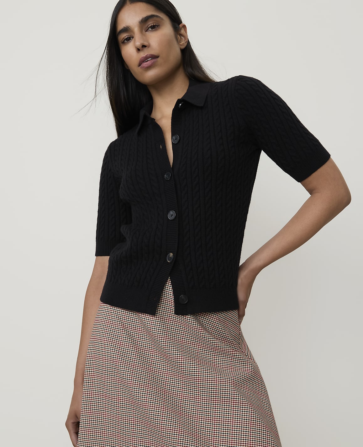 A woman in a black cable-knit polo shirt and a plaid skirt stands against a plain white background.