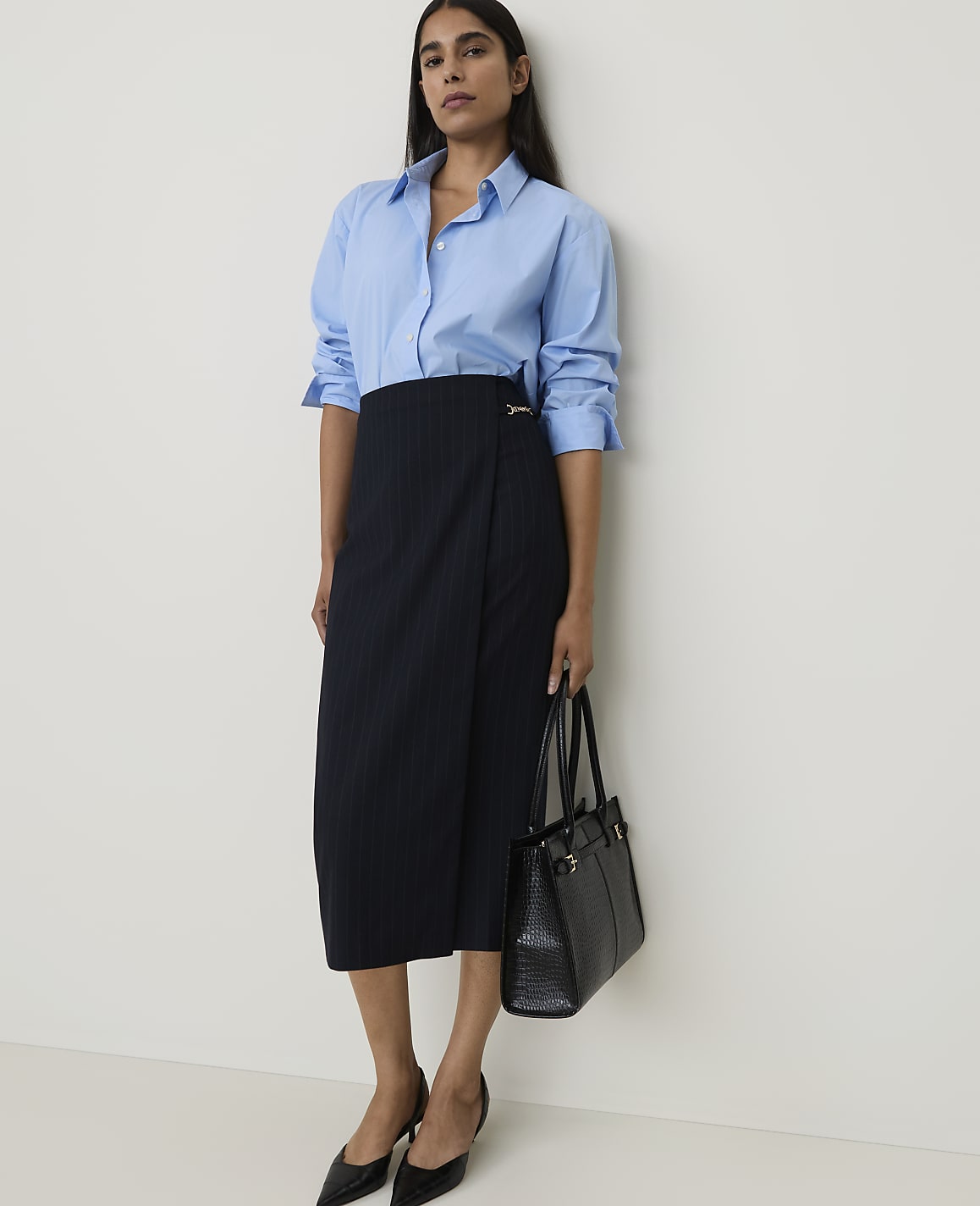 A woman in a blue button-down shirt and pinstripe skirt stands against a plain white wall, holding a black handbag.