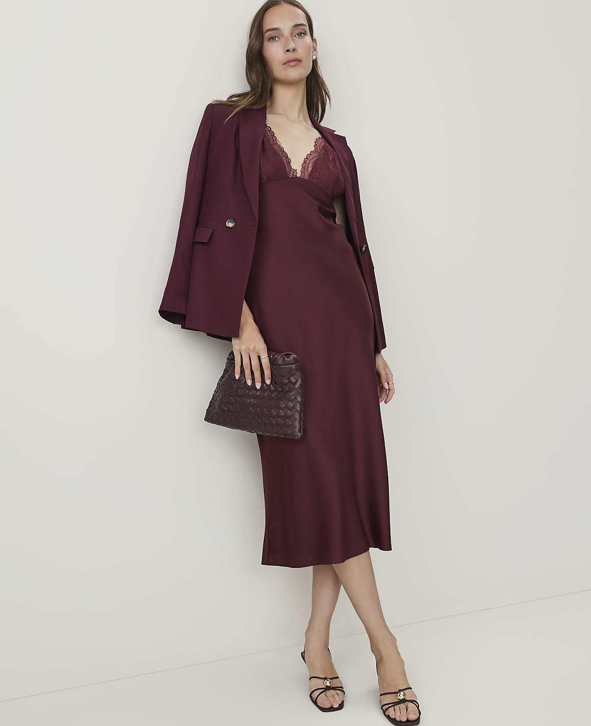 A woman in a burgundy slip dress and blazer holds a woven clutch in front of a plain, light-colored wall.