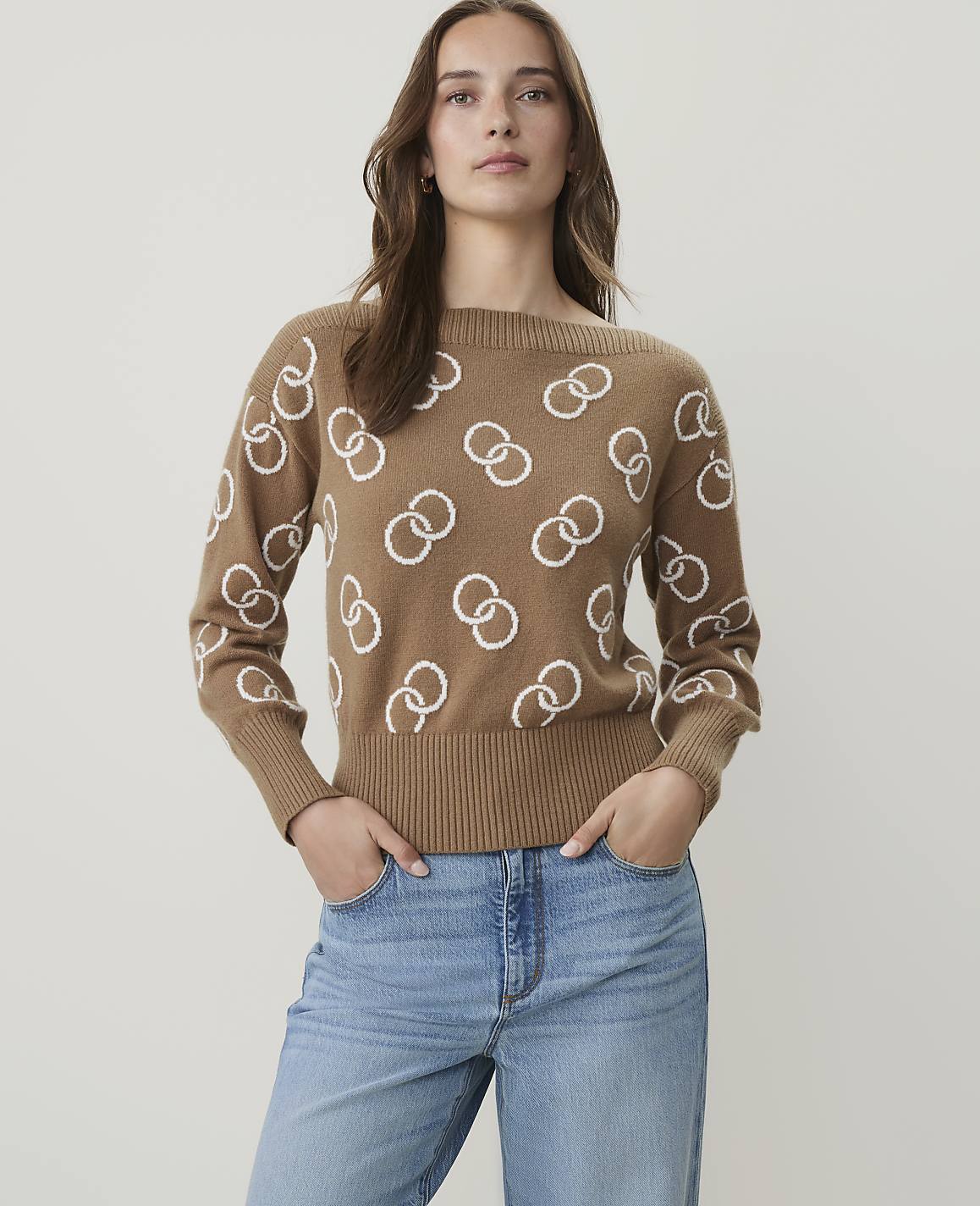 A woman in a tan sweater with a white interlocking ring pattern stands against a plain white background.
