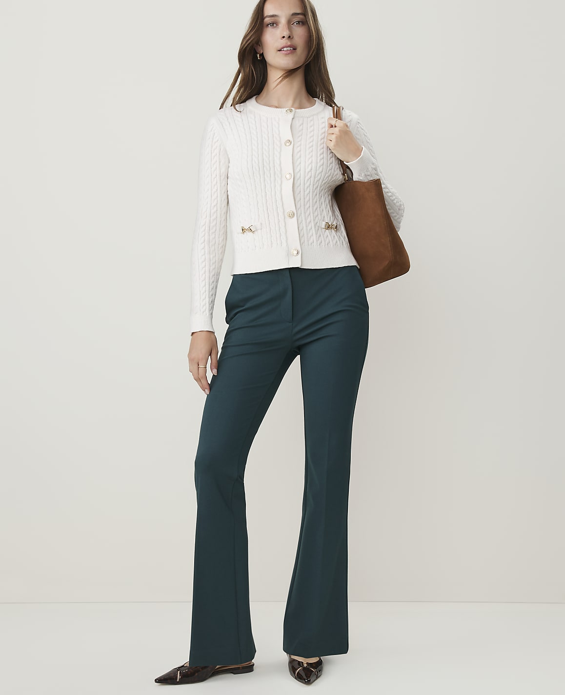 A woman in a white cardigan and teal flared pants stands against a plain, light background, carrying a brown tote bag.