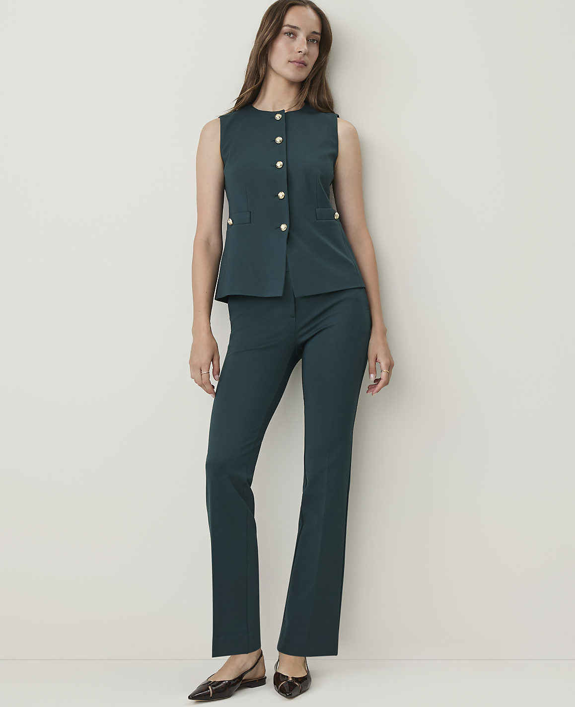 A woman in a teal vest and matching pants stands against a plain, light-colored wall.