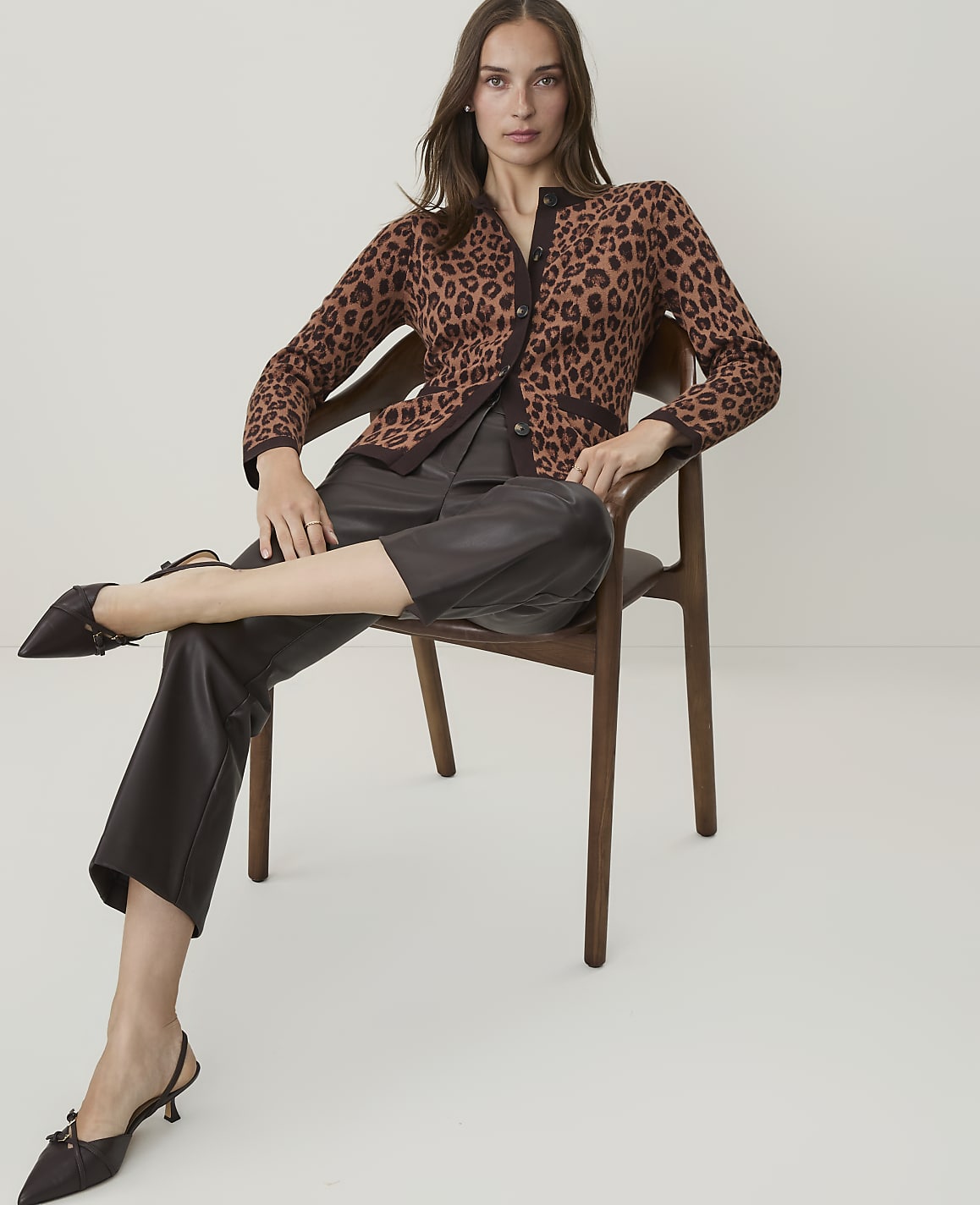 A woman in a leopard print cardigan and dark pants sits on a wooden chair against a plain white background.