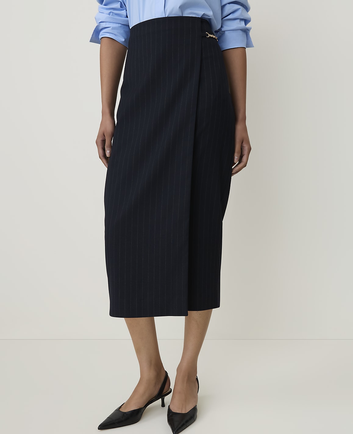 A person stands in a studio setting, wearing a pinstripe wrap skirt and a blue button-down shirt, with black slingback heels on their feet.