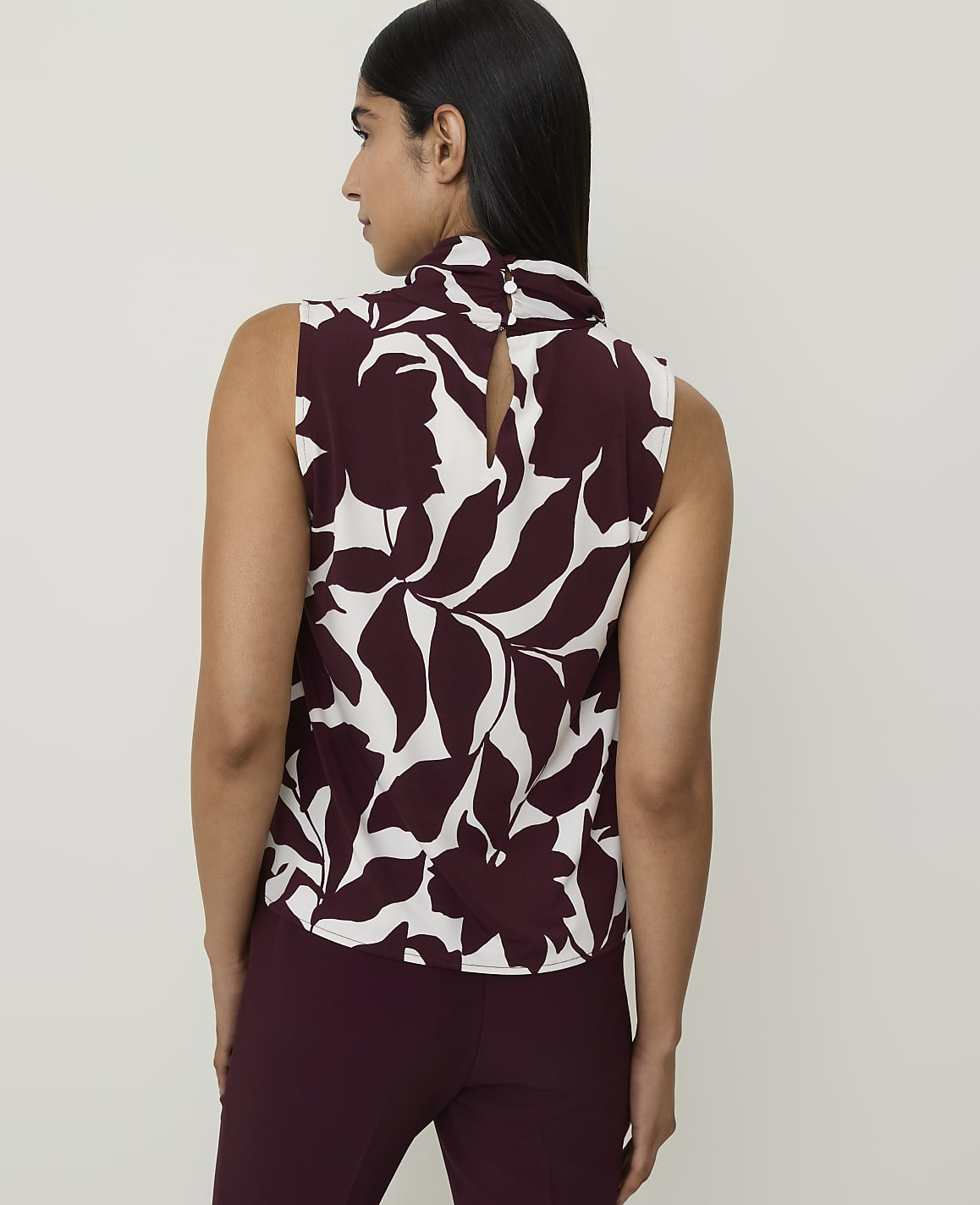 A woman in a floral sleeveless top and maroon pants stands against a plain white background.