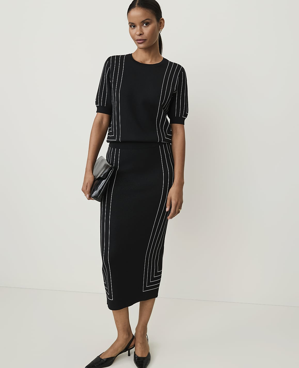 A woman in a black knit set with white piping stands against a plain white background, holding a clutch.