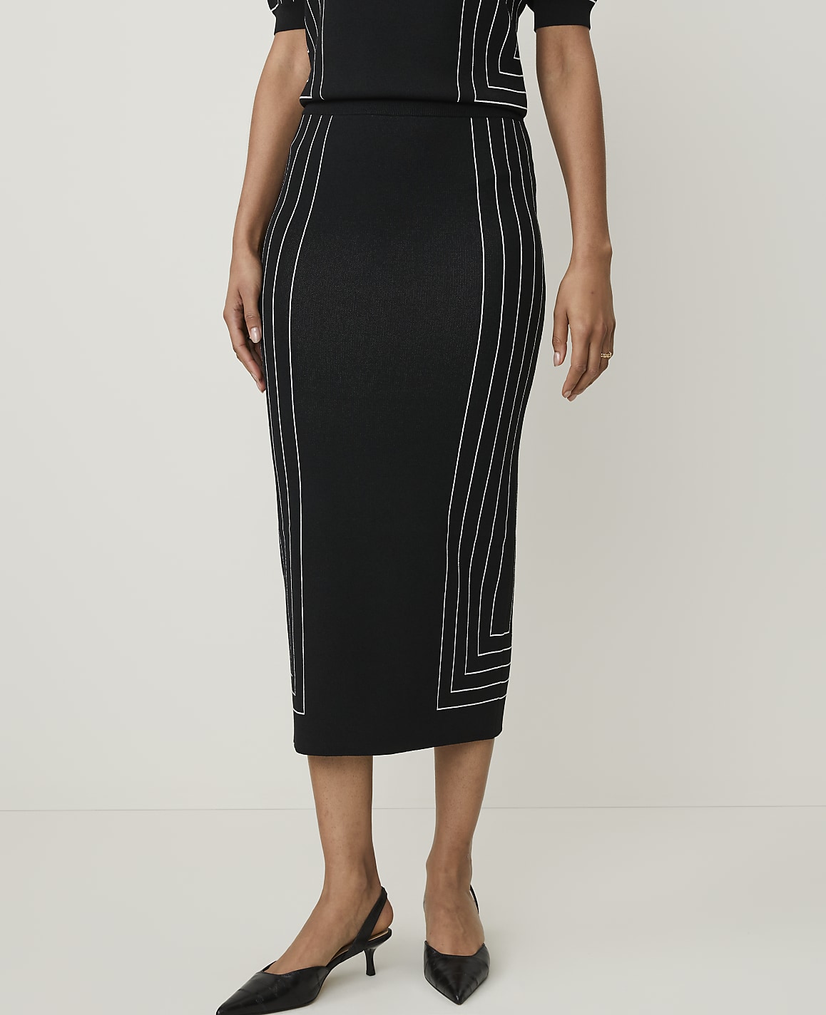 A woman in a black midi skirt with white geometric stitching stands against a plain white background.