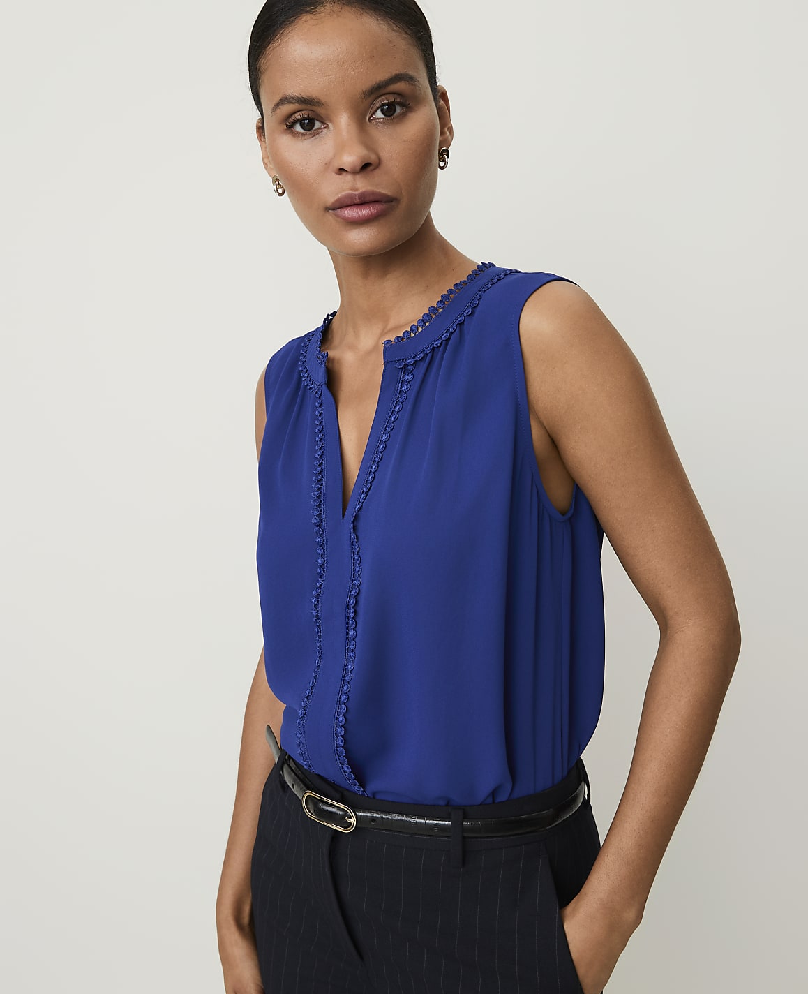 A woman in a blue sleeveless top and pinstripe pants stands against a plain white background.