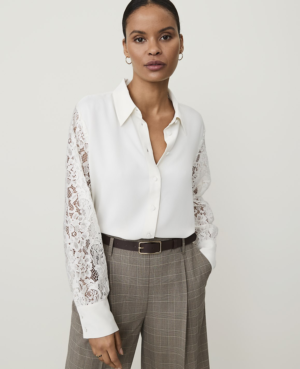 A woman in a white lace-sleeved blouse and plaid trousers stands against a plain, light background.