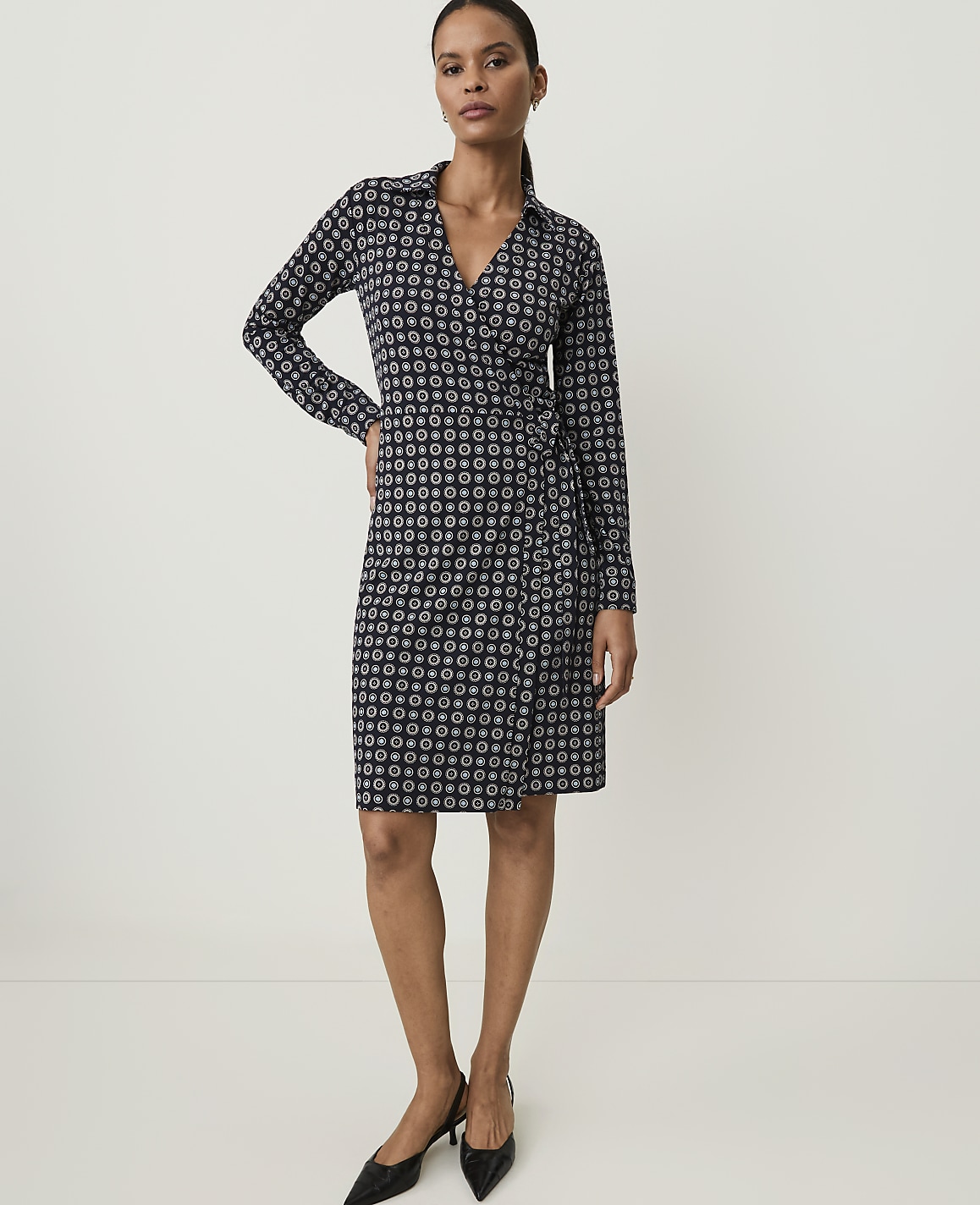 A woman in a patterned wrap dress stands against a plain, light-colored background.