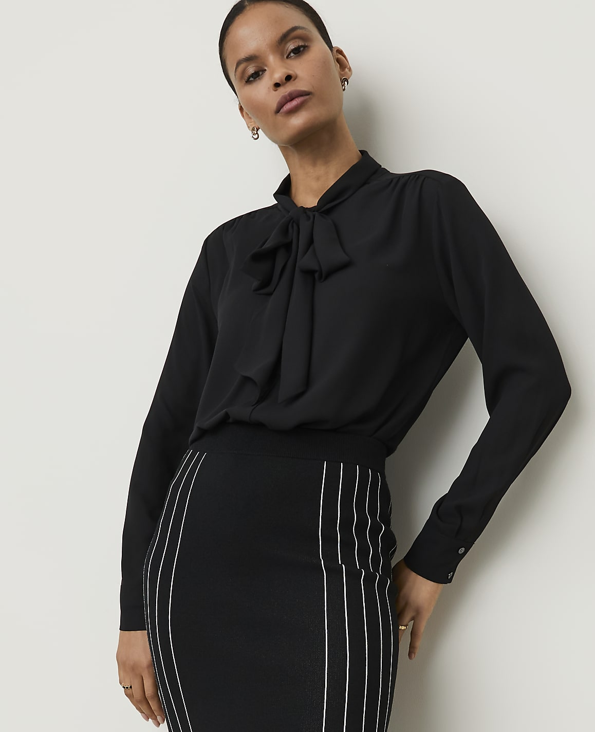 A woman in a black pussy-bow blouse and a striped pencil skirt stands against a plain white background.