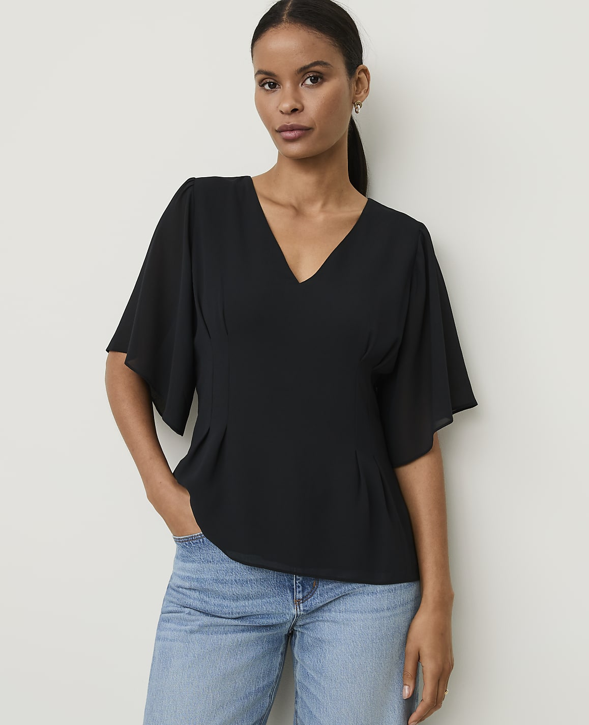 A woman in a black v-neck top and blue jeans stands against a plain white background.
