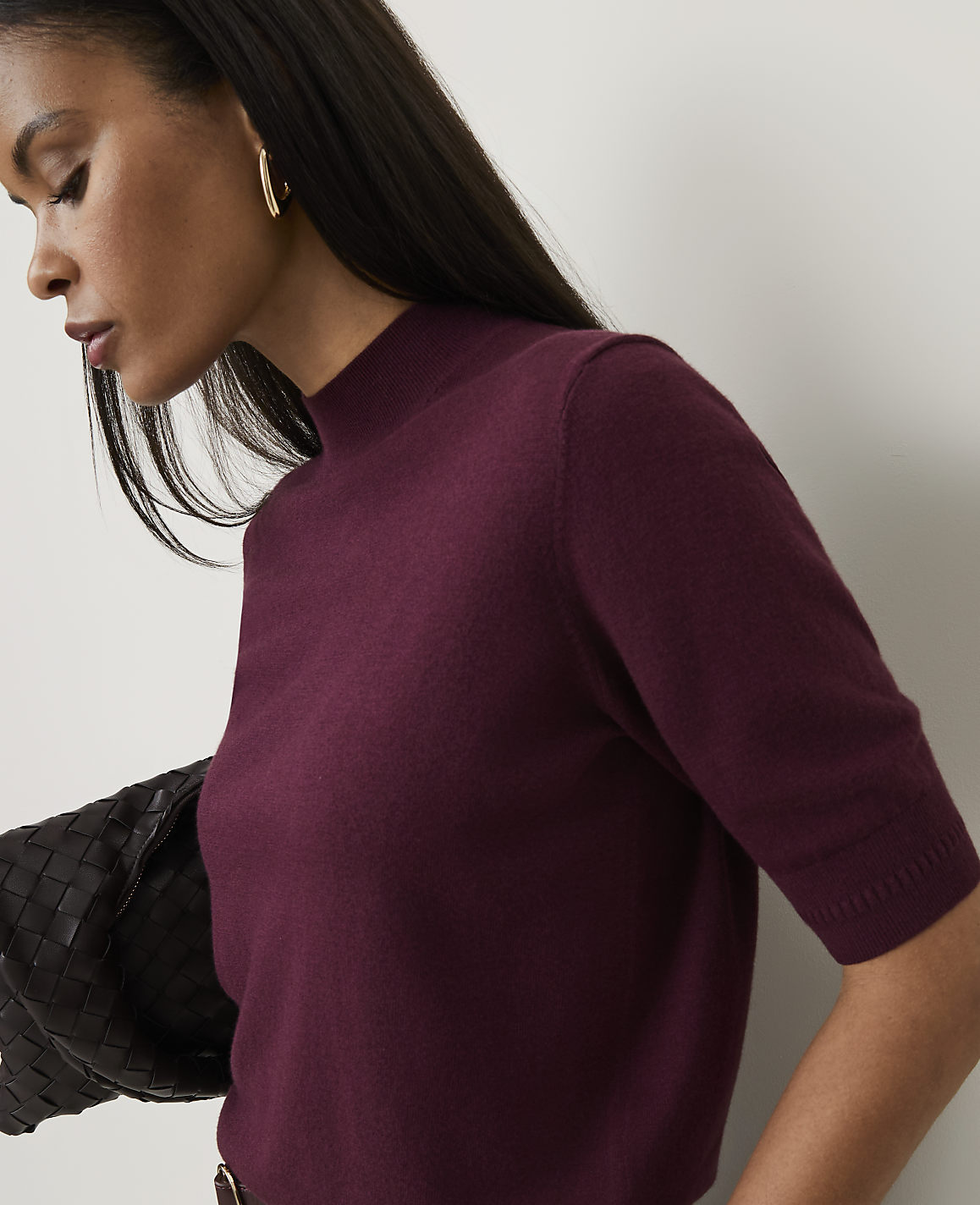 A woman in a burgundy mock-neck sweater is shown in the foreground, with a dark woven bag over her shoulder, against a plain white background.