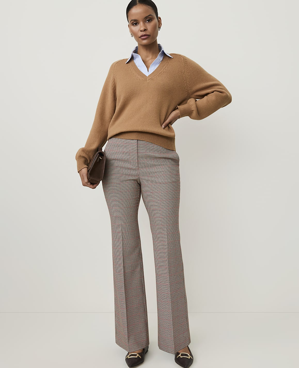 A woman in a camel v-neck sweater, light blue collared shirt, and plaid flared pants stands against a plain, light background, holding a brown clutch.