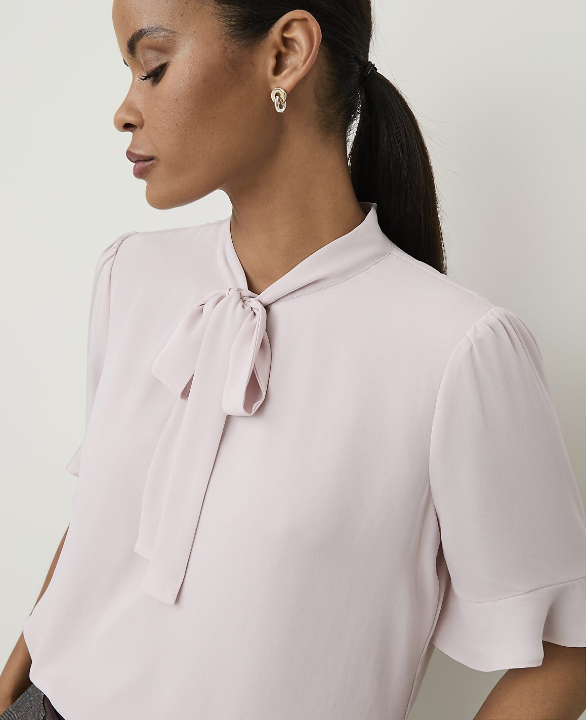 A woman in a pale pink bow-neck blouse with ruffled sleeves is shown in profile against a plain, light background.