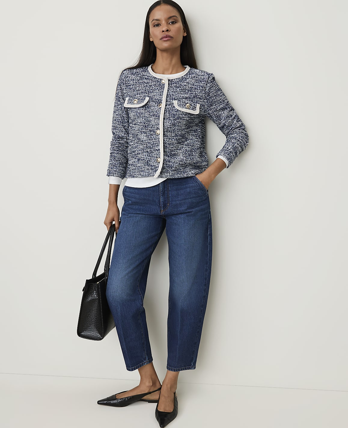 A woman in a tweed jacket and jeans stands against a plain white background, holding a black tote bag.