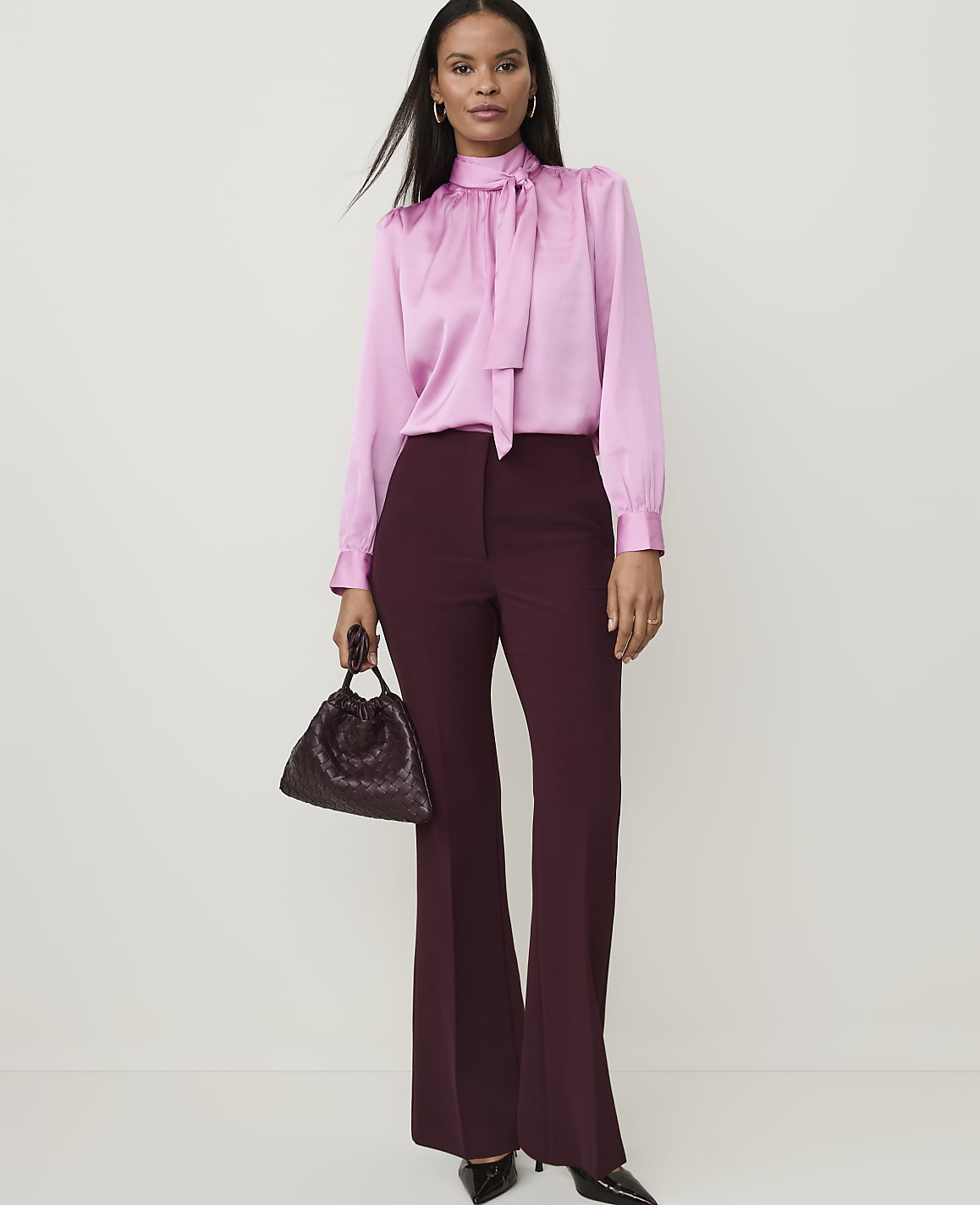 A woman in a pink satin blouse and burgundy flared pants stands against a plain white background, holding a dark, textured handbag.