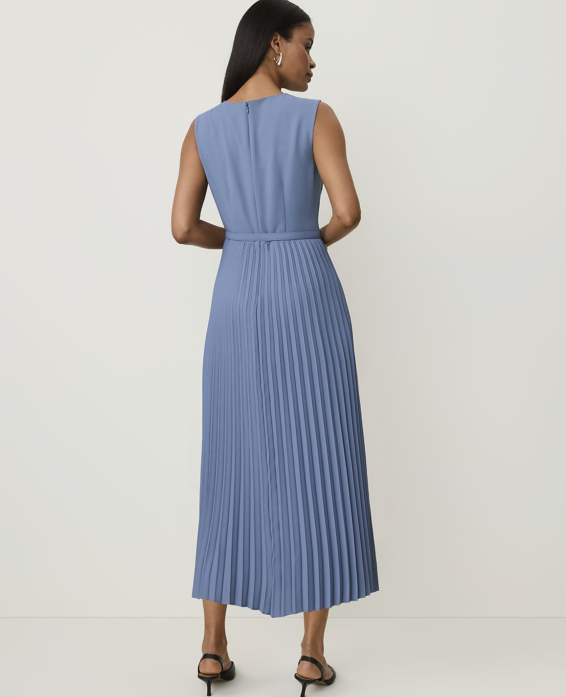 A woman in a blue pleated dress stands against a plain, light-colored background.