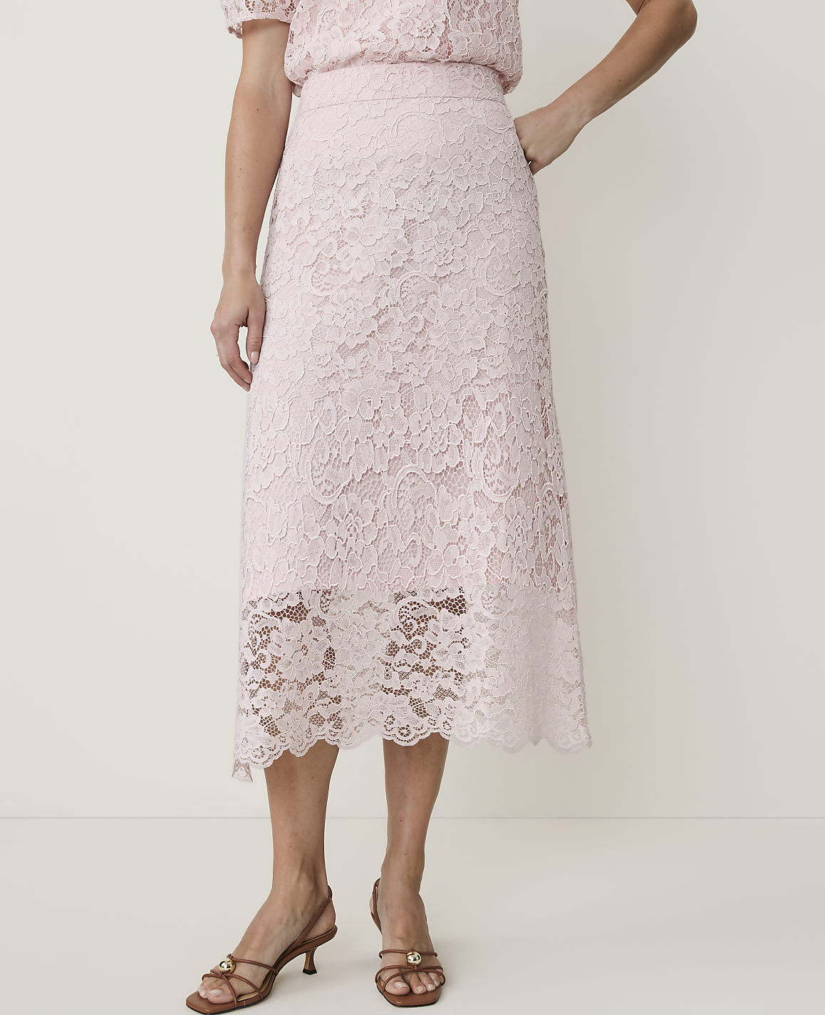 A woman in a light pink lace skirt and matching top stands against a plain white background, her feet adorned with brown heeled sandals.