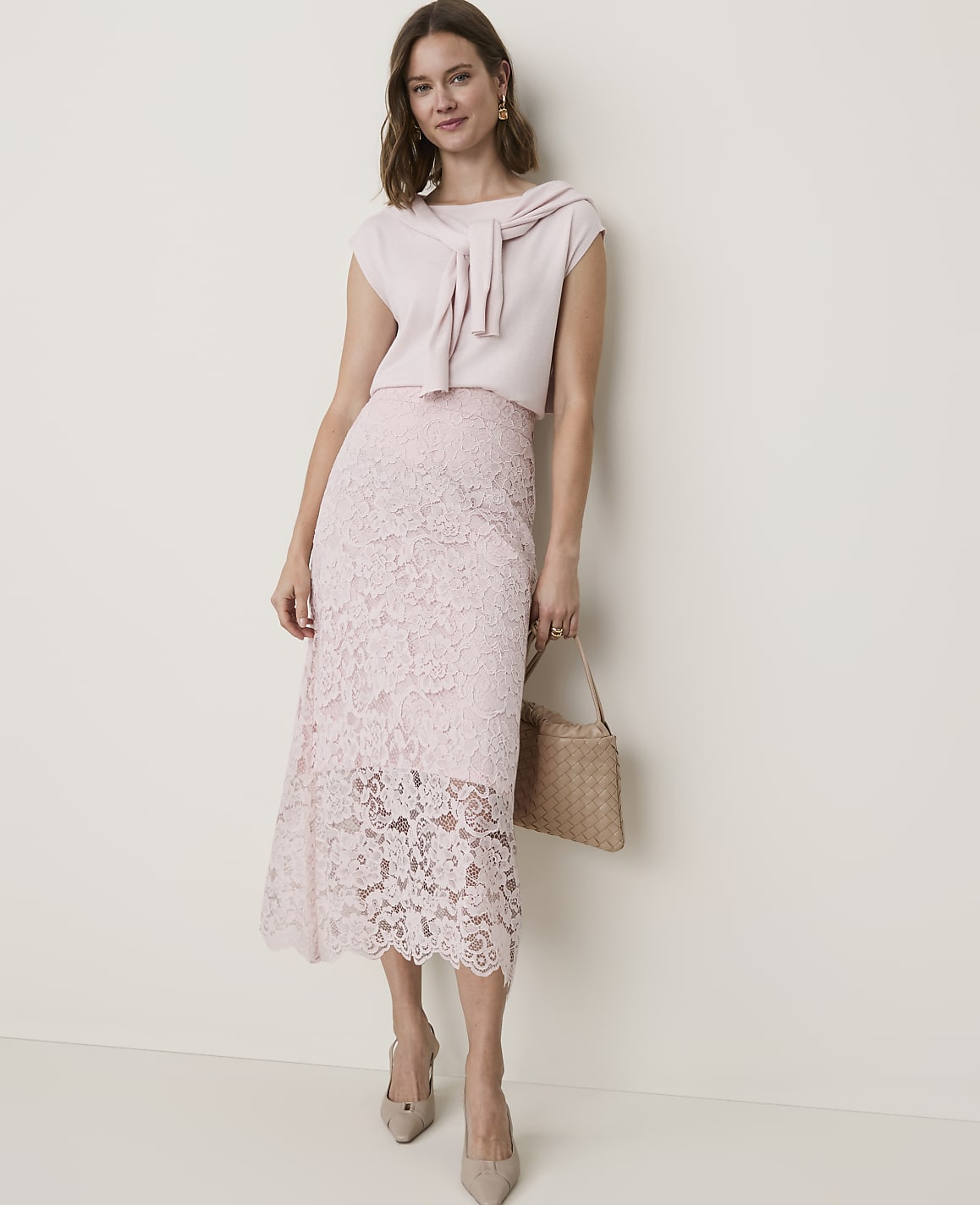 A woman in a light pink lace skirt and matching top stands against a plain, light-colored wall, holding a woven handbag.