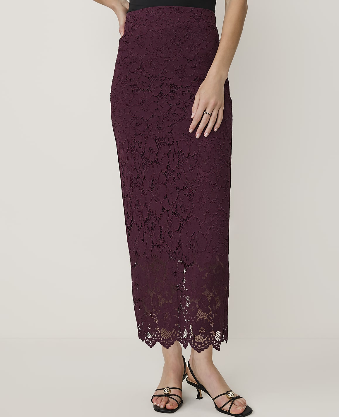 A woman in a burgundy lace pencil skirt and black heels stands against a plain white background.