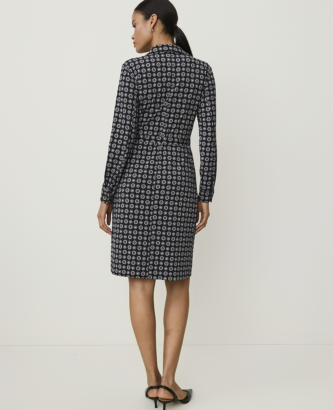 A woman in a patterned dress stands against a plain, light-colored background.