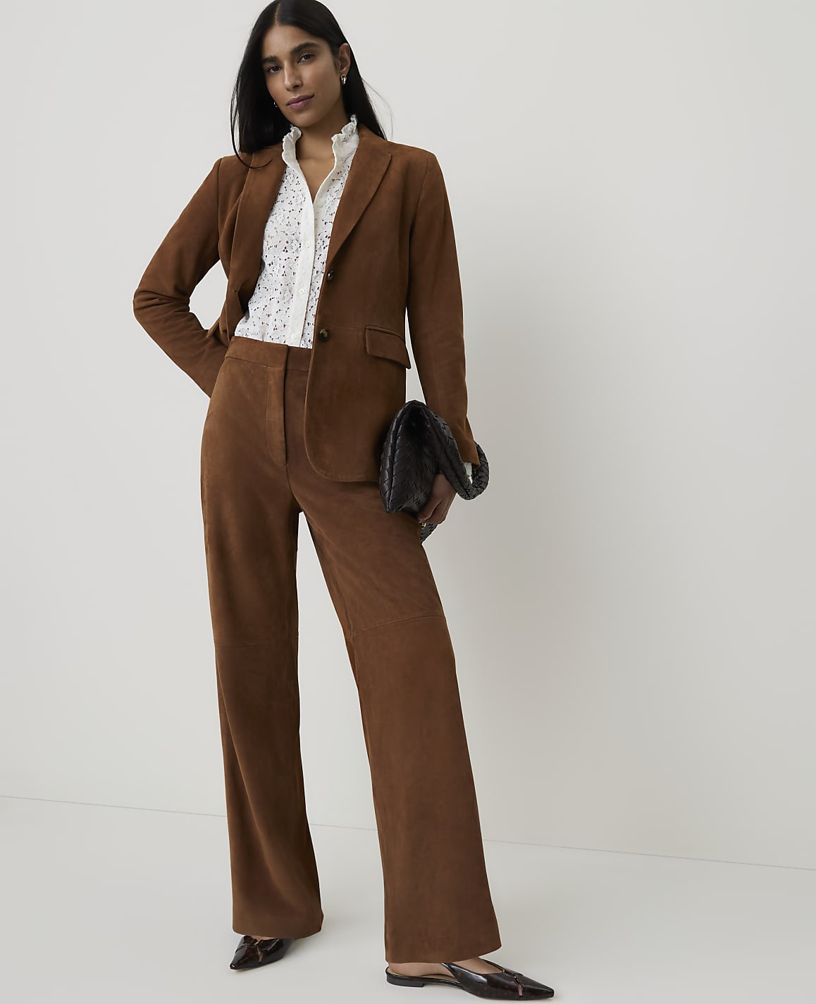 A woman in a brown suit and white blouse stands against a plain white background, holding a black clutch.