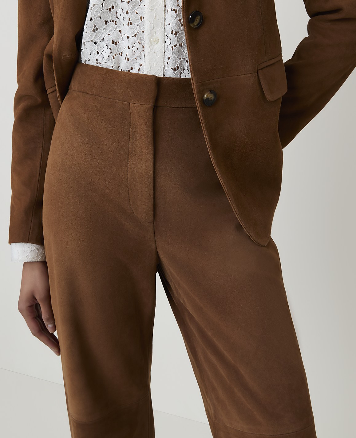 A person wears a brown suede suit with a white lace top, against a plain background.