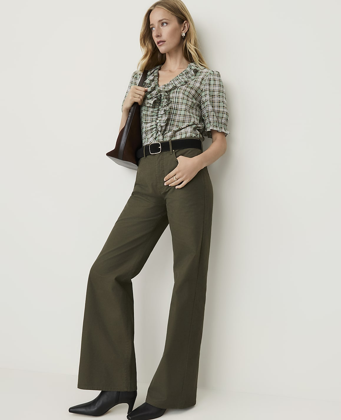 A woman in a green plaid blouse and olive green flared pants stands against a plain white background, holding a brown purse.