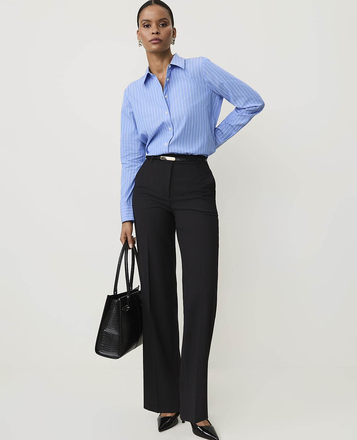 A woman in a blue striped shirt and black pants holds a black handbag against a plain white background.