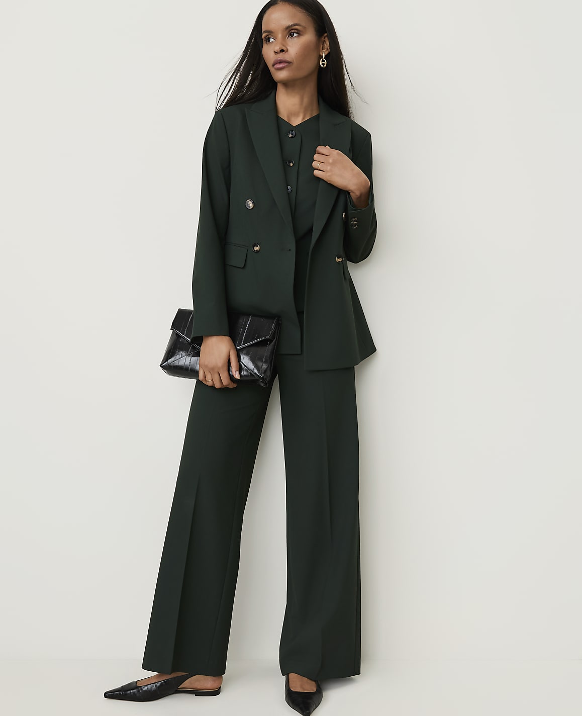 A woman in a dark green pantsuit and holding a black clutch stands against a plain white background.