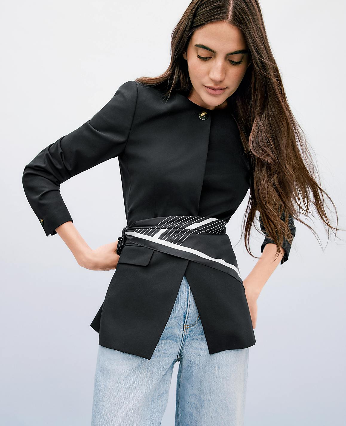 A woman in a black jacket and jeans stands against a light blue background.