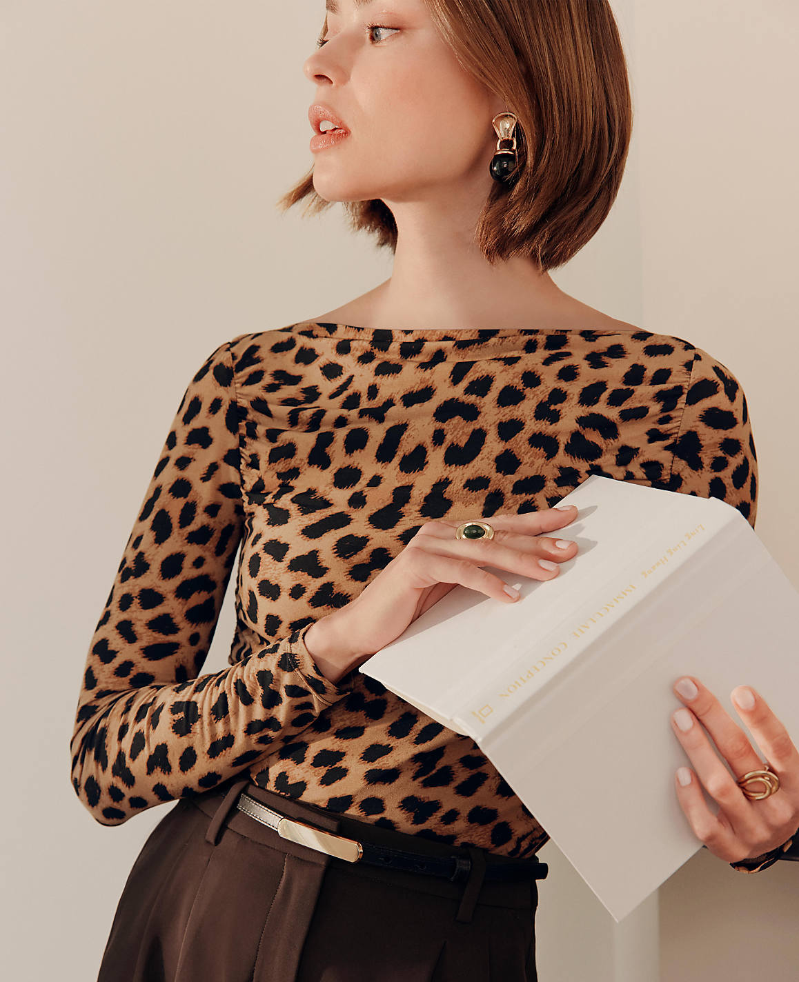 A woman in a leopard print top and brown pants holds a notebook against a plain background.